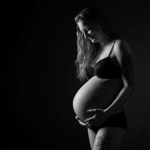 Sort-hvid nærbillede af gravid kvinde med tatoveret arm og sort lingeri i profil, blød belysning mod mørk baggrund
