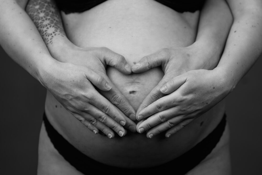 Sort-hvid nærbillede af gravid mave med to par hænder der former et hjerte, tatoveret arm med mandaladesign