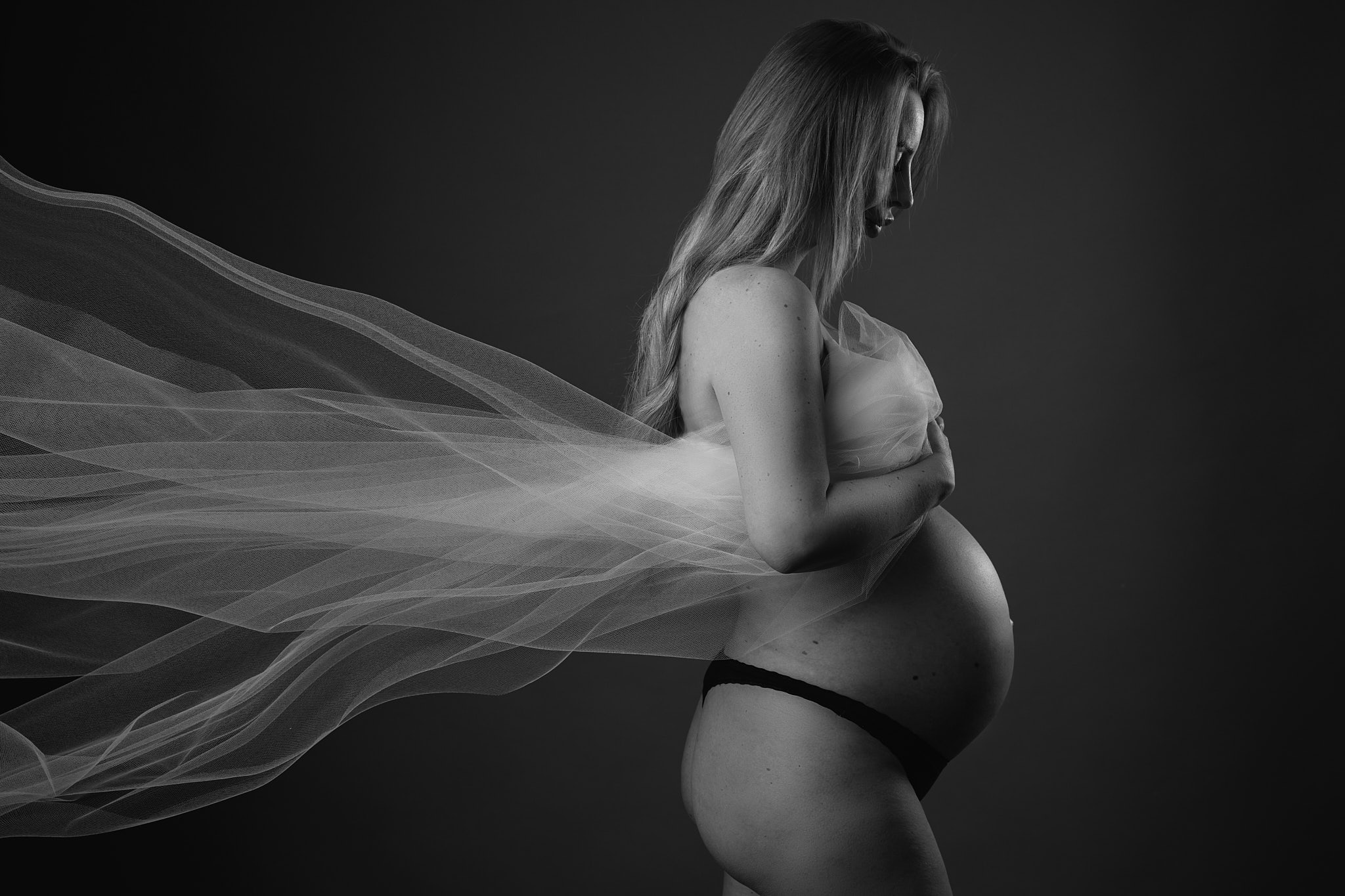 Sort-hvid graviditetsfoto af gravid kvinde med blondt hår i sort lingeri, hvidt slør flyver til siden i profil