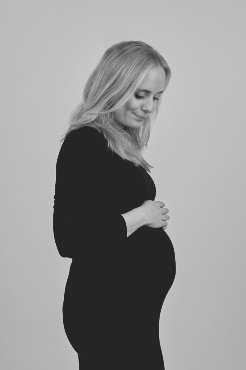 Sort-hvid graviditetsfoto af kvinde i lang sort kjole i profil med langt blondt hår mod lysegrå baggrund