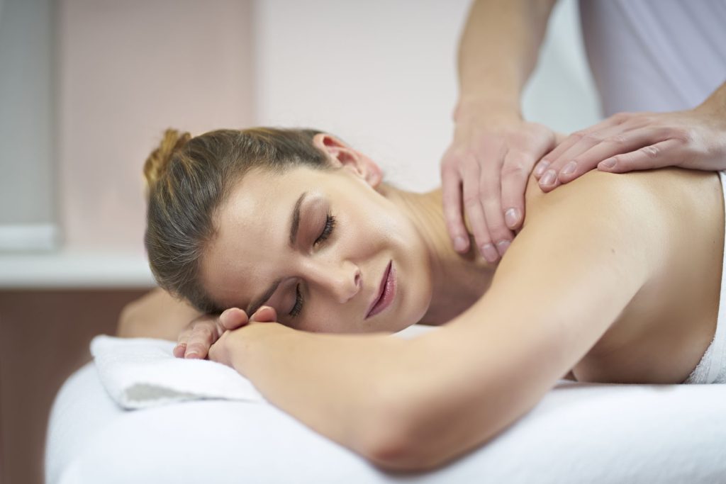 Kvinde på behandlingsbrik modtager skulder- og rygmassage, afslappet udtryk, blødt varmt lys