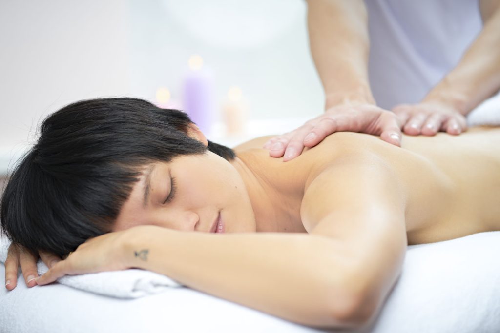 Kvinde med kort mørkt hår modtager rygmassage, stearinlys og lilla lys i blød baggrund