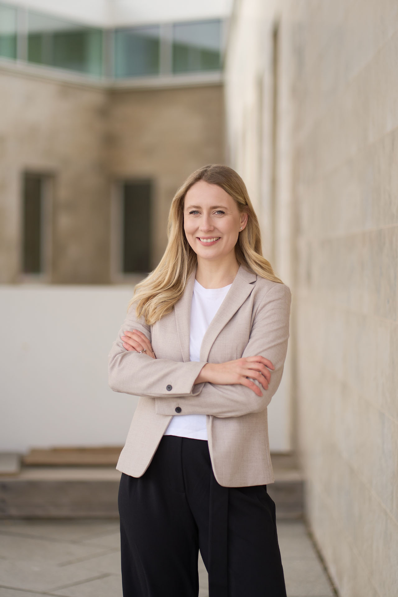 Kvinde i beige blazer med armene over kors smiler bredt foran moderne glasfacader i en udendørs erhvervslokation