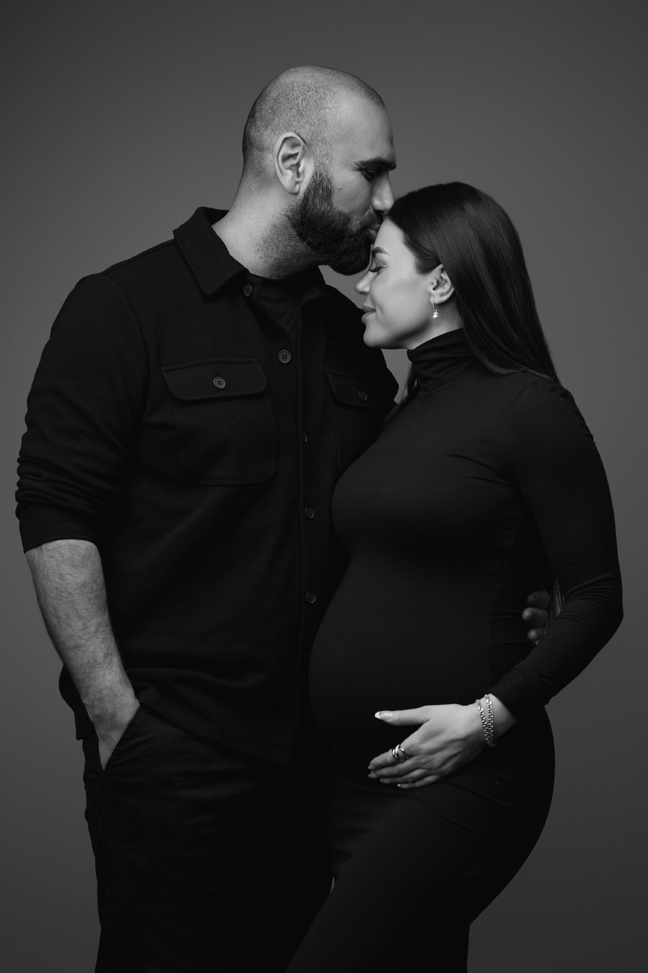 Sort-hvid graviditetsfoto hvor skaldet mand med skæg kysser gravid kvinde i sort turtleneck på panden mens hun smiler