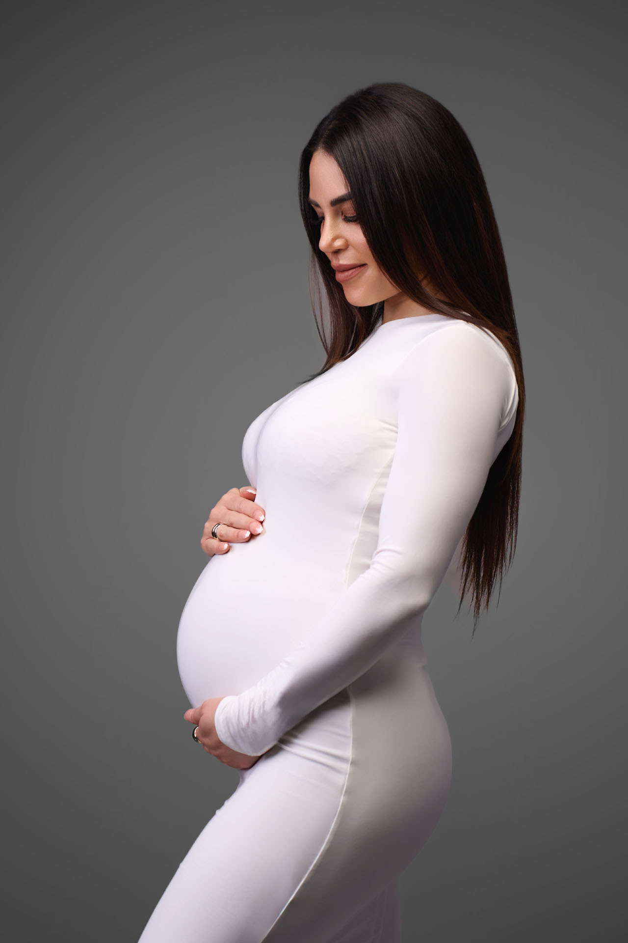 Gravid kvinde i hvid tætsiddende bodysuit med langt sort hår i profil holder om maven mod en grå studiobaggrund