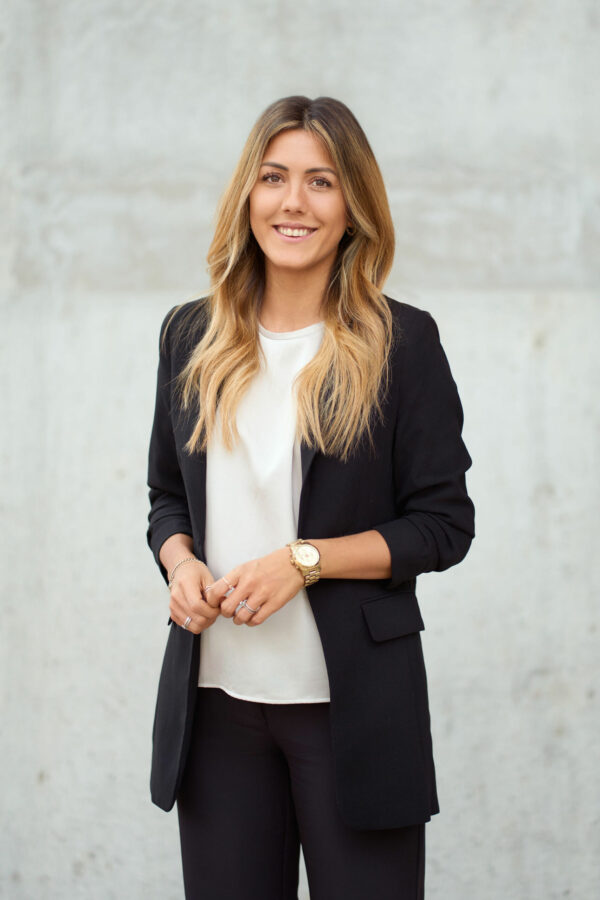 Kvinde med blond ombre hår i sort blazer og hvid skjorte smiler selvsikkert foran grå betonvæg, udendørs erhvervsportræt
