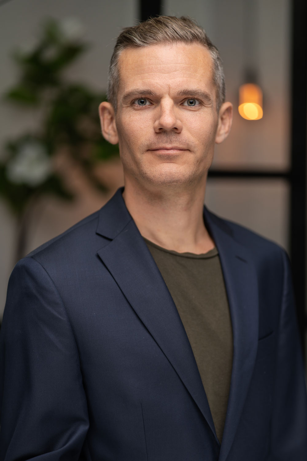 Fotograf René Beck i mørkeblå blazer og olivengrøn t-shirt i halvkropportræt med plante og varm lampe i baggrunden