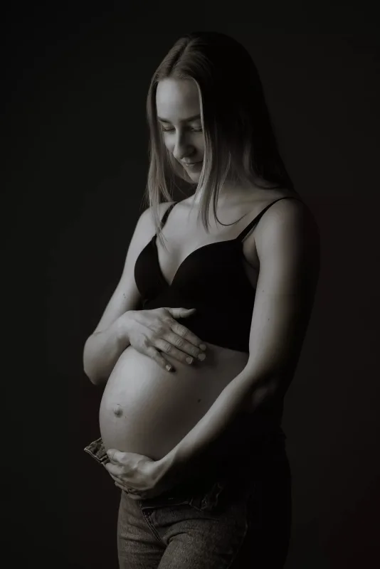 Graviditetsfoto i sort-hvid, gravid kvinde i sort BH holder om maven fra siden, blød studiobaggrund