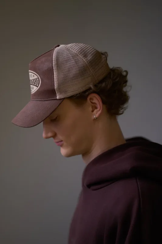 Nærportræt af ung mand i profil med brun trucker cap fra Goodso Copenhagen og mørk bordeaux hoodie. Han kigger ned med lukkede øjne. Dramatisk, mørk studibelysning fremhæver kasketmærket.