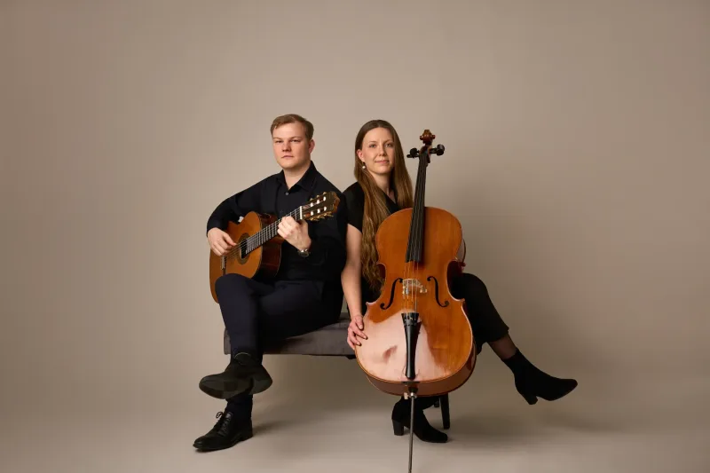 Jonas og Monica i helkropsportræt — han med klassisk guitar, hun med cello — begge kigger direkte i kameraet med et roligt, koncentreret udtryk. Mod varm beige baggrund.
