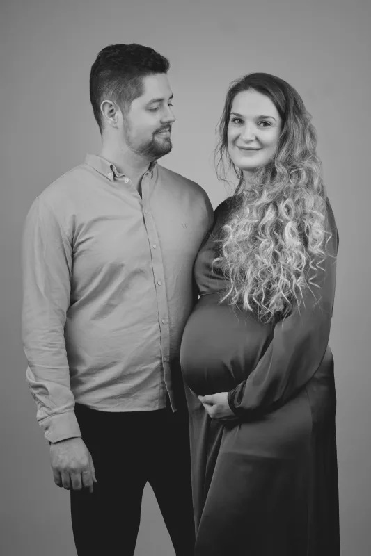 Graviditetsfoto af par i sort-hvid, mand i grå skjorte holder om gravid kvinde med krøllet hår, studiooptagelse