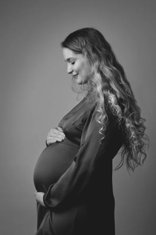 Graviditetsfoto i sort-hvid, gravid kvinde i mørk kjole set fra siden med langt krøllet hår og maven fri i blødt studiolys