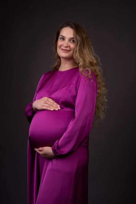Graviditetsfoto af gravid kvinde i lilla satinkjole, hænder foldet over maven, langt krøllet hår og mørk studiobaggrund