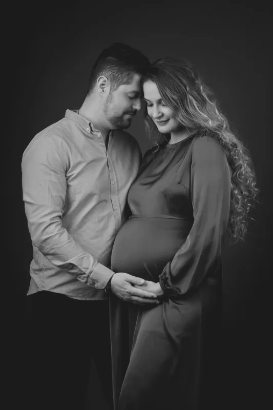 Graviditetsfoto i sort-hvid, smilende par tæt mod hinanden, gravid kvinde med langt krøllet hår og mørk baggrund