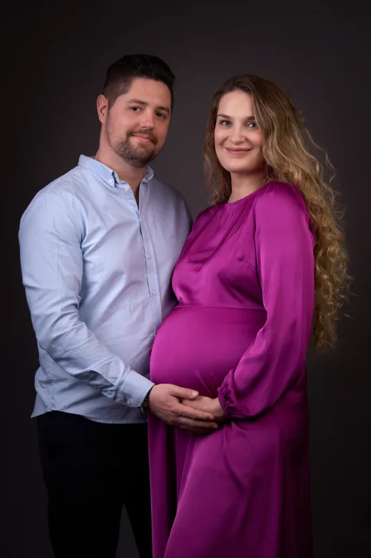 Graviditetsfoto af par i farve, mand i lyseblå skjorte og gravid kvinde i lilla satinkjole, begge vendt mod kameraet