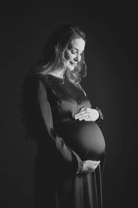 Graviditetsfoto i sort-hvid, gravid kvinde i mørk kjole fra siden, maven fremhævet, langt krøllet hår i dramatisk sidelys