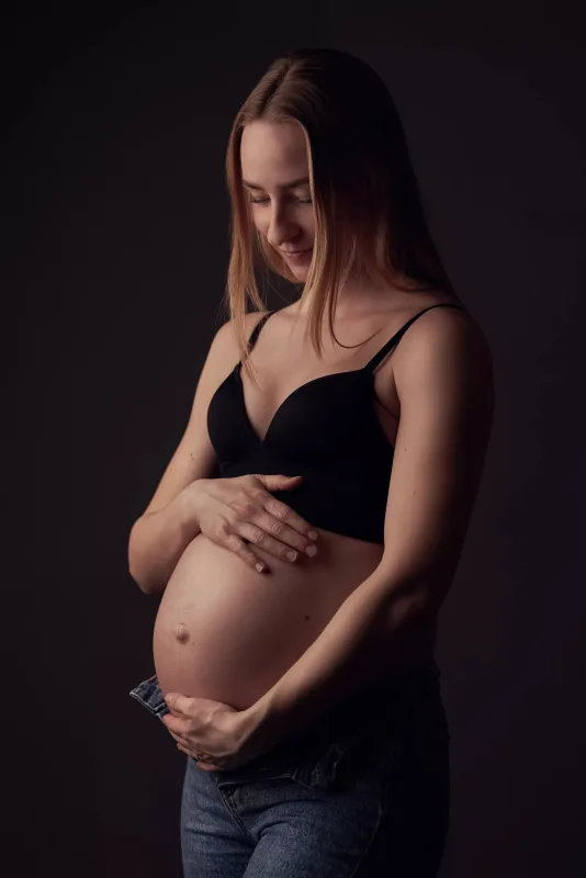 Graviditetsfoto, gravid kvinde i sort blonde-BH holder om den runde mave, set fra siden med mørk studiobaggrund