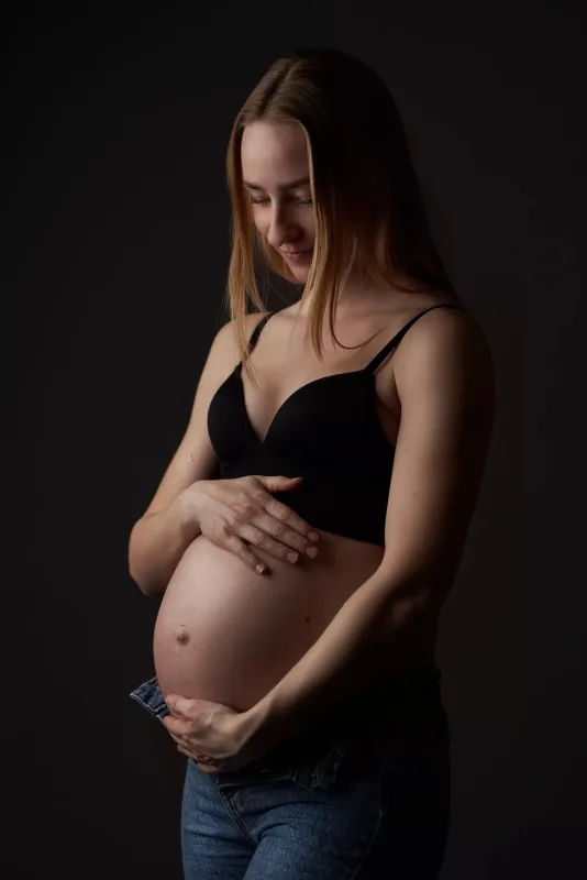 Graviditetsfoto, gravid kvinde i sort BH med hænder om maven, let sidelys fremhæver runde mave mod mørk studiobaggrund
