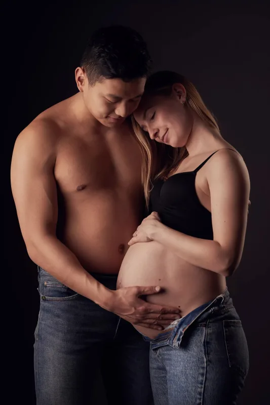 Graviditetsfoto af par, topless mand holder om gravid kvinde i sort BH, begge kigger ømt ned mod maven, mørk baggrund
