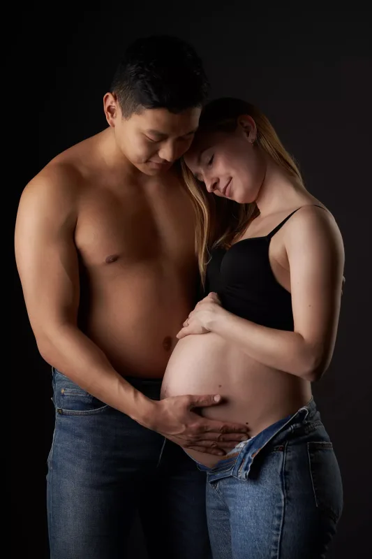 Graviditetsfoto af par, topless mand og gravid kvinde i sort BH tæt på hinanden, intim stemning foran mørk studiobaggrund