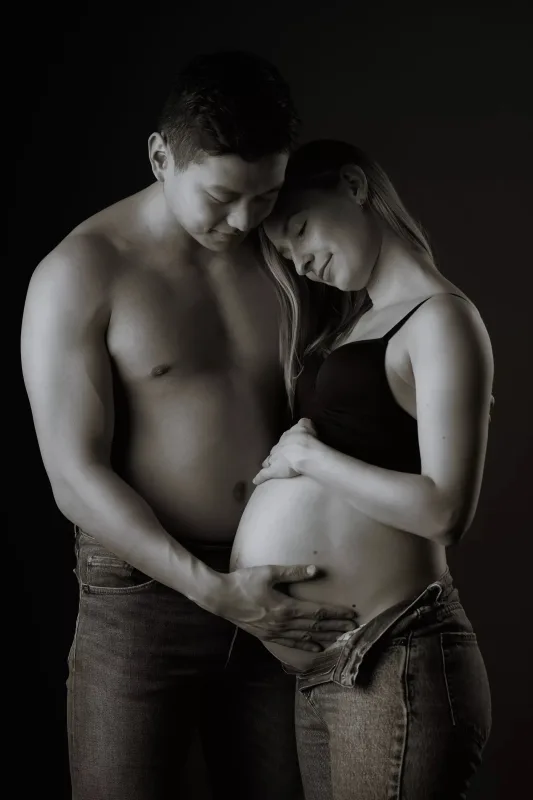 Graviditetsfoto i sort-hvid, topless mand holder om gravid kvinde i sort BH, beskyttende positur foran mørk baggrund