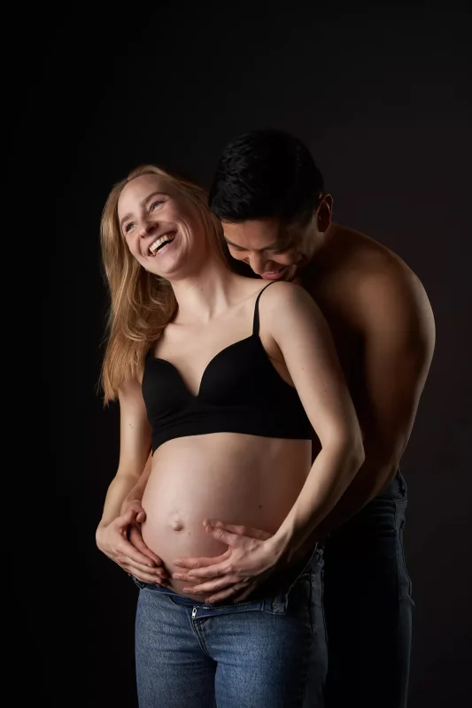 Graviditetsfoto af smilende par, gravid kvinde med langt blondt hår i sort BH og kærlig mand i lysende studiomiljø