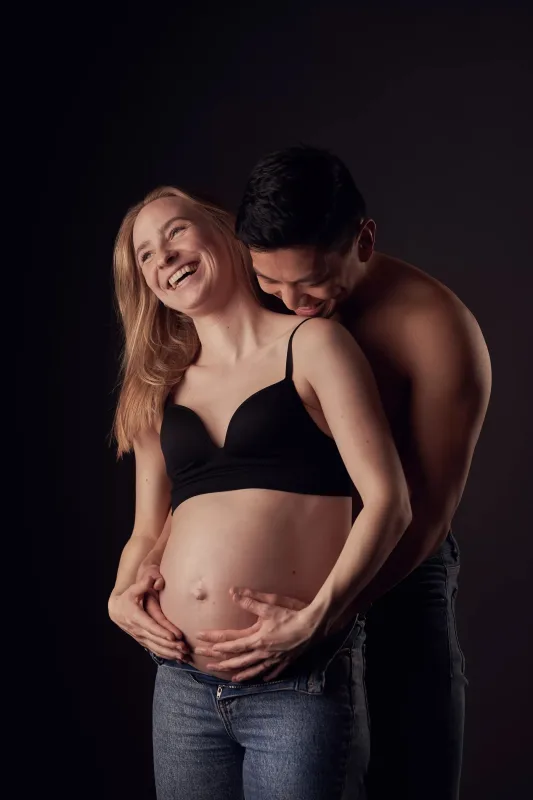 Graviditetsfoto med grinende par, gravid kvinde i sort BH og langt blondt hår i glædesfyldt øjeblik i studie