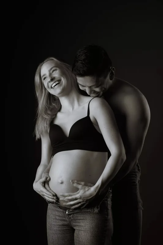 Graviditetsfoto i sort-hvid, smilende par i studie, gravid kvinde med langt hår i sort BH og manden tæt ved