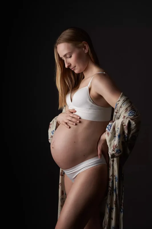 Graviditetsfoto af gravid kvinde i hvid sports-BH med åben blomstret jakke, maven fremhævet fra siden, mørk baggrund