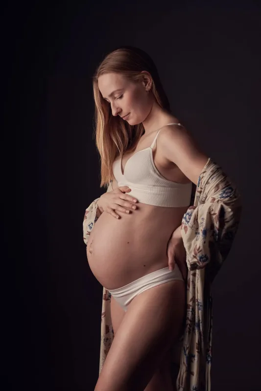 Graviditetsfoto, gravid kvinde i hvid BH og blomstret åben jakke holder forsigtigt om maven fra siden, blød studiabelysning