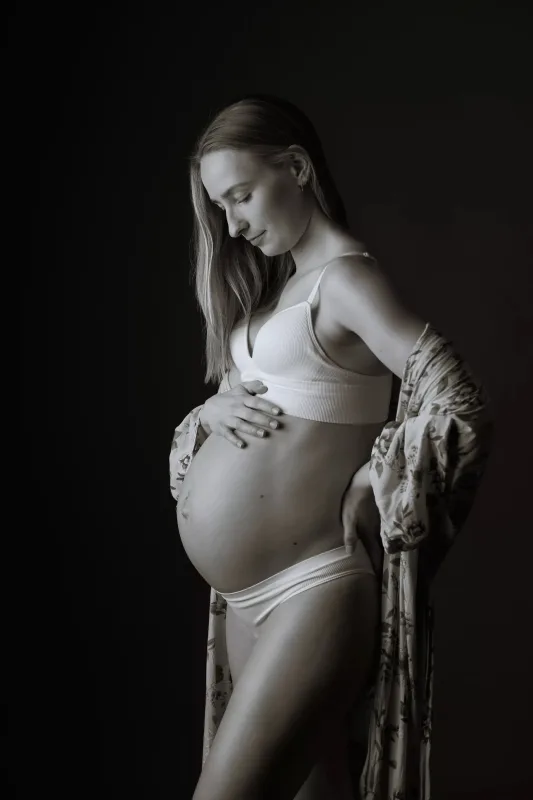 Graviditetsfoto i sort-hvid af gravid kvinde i hvid BH med åben blomstret jakke, maven fremhævet fra siden i studiomiljø